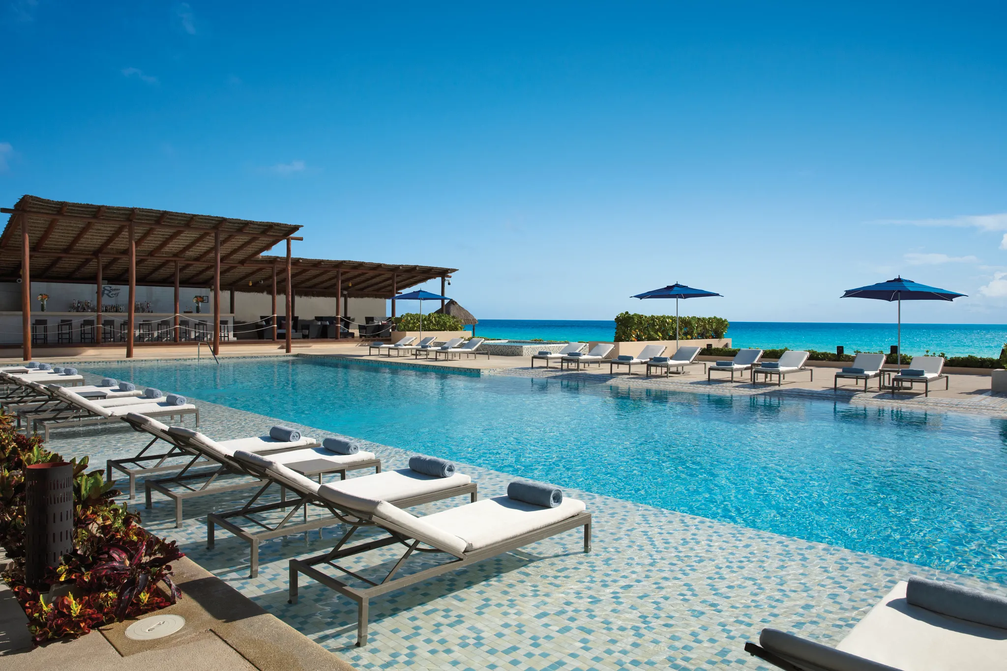 Infinity pool with lounge chairs and oceanfront bar at Secrets The Vine Cancun under a clear blue sky.